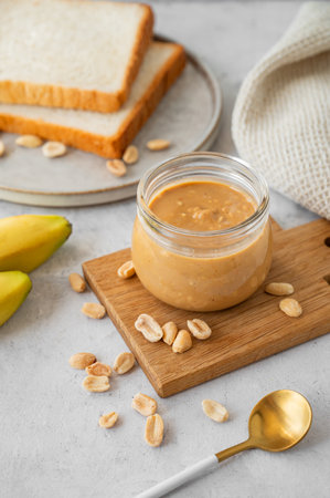Glass jar filled with creamy peanut butter on grey background with bananas and fresh bread. Natural homemade nut butter concept.の写真素材