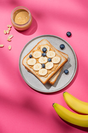 Flat lay of peanut butter toast with banana, blueberry and nuts on a plate on a pink background with shadow. Concept of sweet and healthy snacks.の写真素材