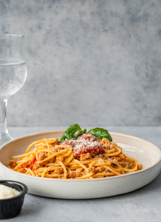 Spaghetti bolognese with tomato sauce, basil and parmesan cheese on a plate on a gray background with glass of water. The concept of Italian or Mediterranean diet cuisine. Copy space.の写真素材