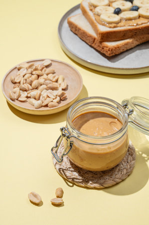 Peanut butter in glass jar on yellow background with salted nuts, toast, banana and berries. Concept of tasty and healthy snack for sweet sandwiches for breakfast.の写真素材