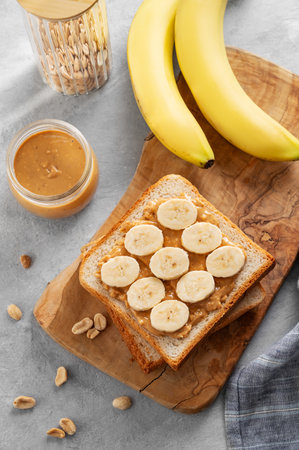 Peanut butter toast with banana on a wooden board on a light background with fruits and morning shadow. Concept of sweet diet and healthy snacks for american breakfast. Top view and copy space.の写真素材