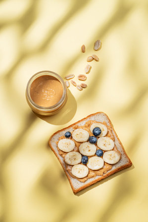 Peanut butter toast with banana, blueberry and nuts on a yellow background with shadow. Concept of...