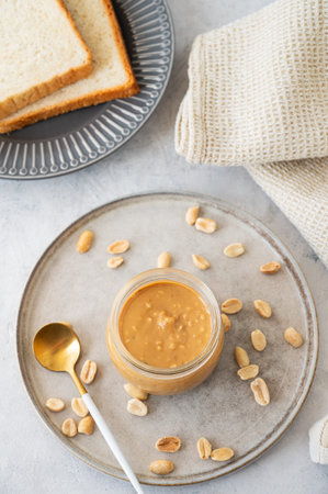 Peanut butter in glass jar on light  background with salted nuts and bread toasts. Concept of tasty...