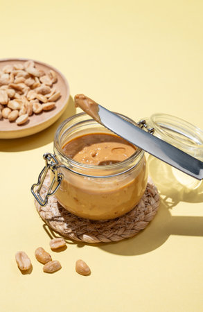 Peanut butter in glass jar on yellow background with knife and salted nuts. Concept of tasty and healthy snack for sweet sandwiches for breakfast. Copy space.の写真素材