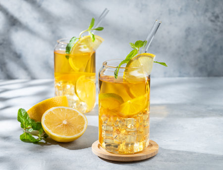 Two glasses with iced drink with lemon and mint on a light background with shadows and citrus fruits. The concept of a cold tea or lemonade on a  summer day. Front view, copy space.の写真素材