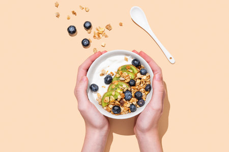Hands holding bowl of natural yogurt with granola, kiwi and blueberry on beige background. Healthy and nutritious breakfast concept, cafe menu advertising banner. Top view, copy spaceの写真素材