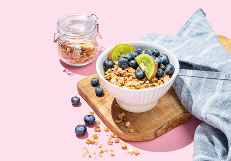 Baked granola or muesli in a bowl with kiwi and blueberries on a wooden board on a pink background. Healthy and creative breakfast concept for food advertising or menu layout. Copy space.の写真素材