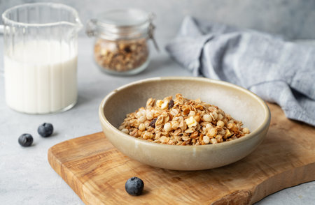 Homemade baked granola or muesli in a bowl on a wooden board on a light background with milk blueberry and napkin close up. Healthy and nutritious breakfast concept. Front  view.の写真素材