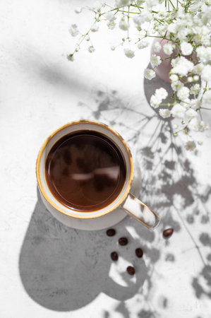 Cup of freshly brewed coffee and bouquet of gypsophila on light textured background with morning shadows and coffee beans. Aesthetic concept of breakfast drink. Top view and copy space.の写真素材