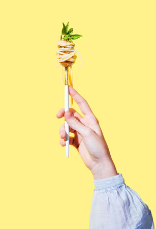 Hand holding fork with creamy spaghetti and basil on isolated bright yellow background. Italian or Mediterranean cuisine concept for menu banner or restaurant advertisement. Front view, copy space.の写真素材