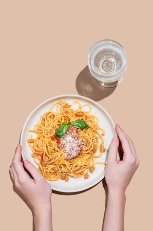 Hands holding a plate of spaghetti bolognese with tomato sauce, basil and cheese on a beige background with a glass of water. Italian cuisine concept for banner advertising of menu. Top view.の写真素材