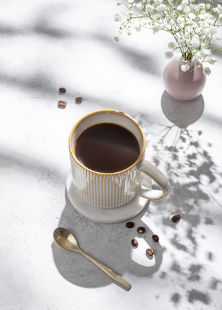 Cup of freshly brewed coffee and bouquet of gypsophila on light textured background with morning shadows and coffee beans. Aesthetic concept of breakfast drink. Top view and copy space.の写真素材