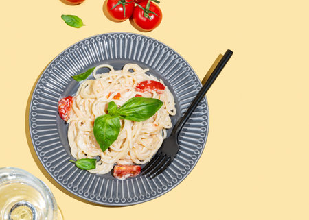 Creamy spaghetti with tomatoes, cheese and basil on a plate on a yellow background with a glass of water. Italian or Mediterranean cuisine concept for menu banner or advertising. Top view, copy spaceの写真素材