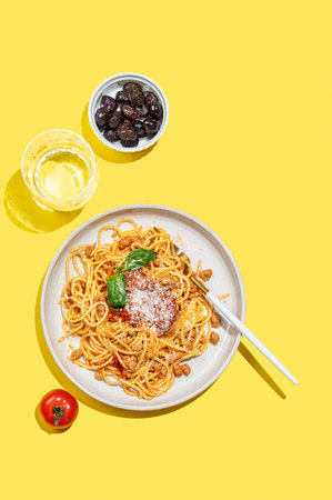 Spaghetti bolognese with tomato sauce, basil and parmesan cheese on yellow background with glass of water and olives. Italian cuisine concept for menu. Top view, copy space.の写真素材