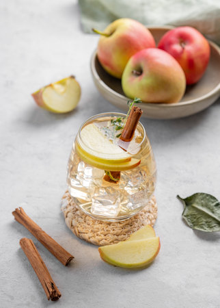 Chilled apple cocktail or cider in a glass with ice, garnished with cinnamon and thyme on a light background with fruits. Refreshing drink concept.の写真素材