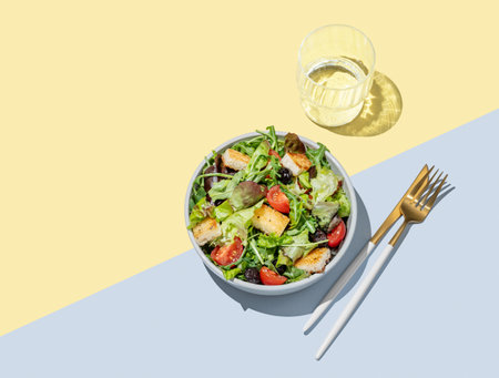 Vegetable salad with lettuce, tomatoes, croutons and olives in a bowl on a yellow blue background with glass of water and cutlery. Healthy diet food concept  for advertising menu. Top view, copy space.の写真素材