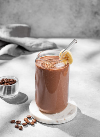 Chocolate coffee smoothie with banana in a glass on a light blue  background with morning shadow. Healthy eating and detox diet concept for advertising menu or banner. Front view, copy space.の写真素材