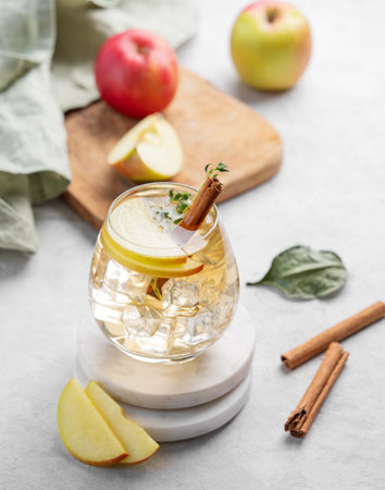 Chilled apple cocktail or cider in a glass with ice, garnished with cinnamon and thyme, on a marble stand on a light background with fruits. Refreshing drink concept.の写真素材