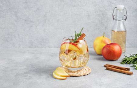 Apple cocktail or cider  with ice, garnished with cinnamon and rosemary  in a glass on a light background with slice of fruits and bottle. Refreshing drink concept. Front view, copy spaceの写真素材