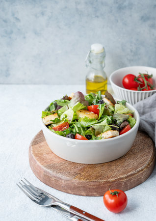 Vegetarian salad with lettuce, tomatoes, croutons and olives in a bowl on a wooden board on a light background with fresh vegetables, oil and cutlery. Healthy diet food concept. Copy space.の写真素材