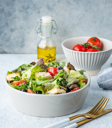 Salad with lettuce, tomatoes, croutons, olives and parmesan cheese in a bowl on a light background with fresh vegetables, oil and cutlery. Healthy diet food concept. Front view, copy space.の写真素材