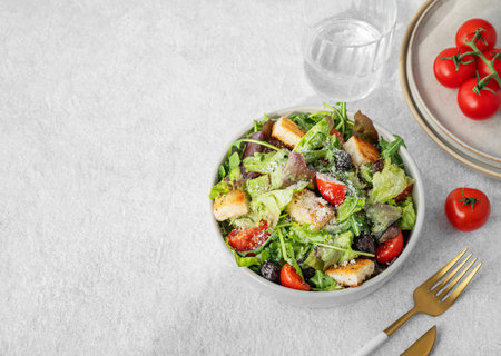 Vegetarian salad with lettuce, tomatoes, croutons, olives and parmesan cheese in a bowl on a light background with fresh vegetables and cutlery. Healthy diet food concept. Top view, copy spaceの写真素材