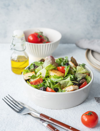 Vegetarian salad with lettuce, tomatoes, croutons and olives in a bowl on a light background with fresh vegetables, oil and cutlery. Healthy diet food concept. Copy space.の写真素材