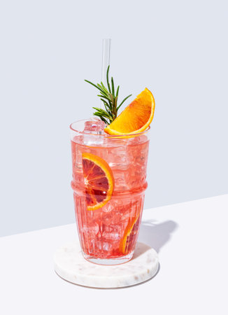Blood orange cocktail with ice and rosemary on a marble stand on a white blue background with shadow. Summer citrus drink concept for cafe menu advertising banner. Front view and copy space.の写真素材