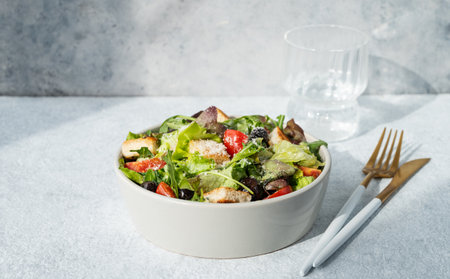 Salad with lettuce, tomatoes, croutons, olives and parmesan cheese in a bowl on a light background with sunlight, glass of water and cutlery. Healthy diet food concept. Front view, copy space.の写真素材