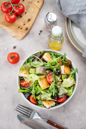 Vegetable salad with lettuce, tomatoes, croutons and olives in a bowl on a light background with spices,   oil and cutlery. Healthy diet food concept. Top view.の写真素材