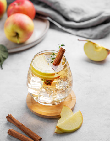 Chilled apple cocktail or cider in a glass with ice, garnished with cinnamon and thyme on a light background with fruits. Refreshing drink concept.の写真素材