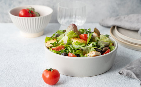 Vegetarian salad with lettuce, tomatoes, croutons and olives in a bowl on a light background with fresh vegetables and glass of water close up. Healthy diet food concept. Front view, copy space.の写真素材