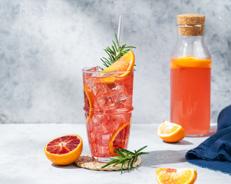 Blood orange cocktail with ice and rosemary on a marble stand on a gray background with bottle and shadows. Summer citrus drink concept for cafe menu. Front view, copy space.の写真素材