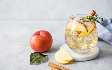 Apple cocktail or cider  with ice, garnished with cinnamon and thyme in a glass  on a marble stand on a light background with slice of fruits. Refreshing drink concept. Front view, copy spaceの写真素材