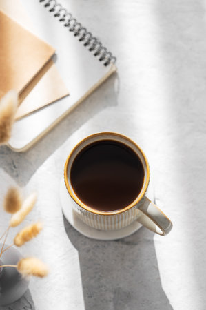 Freshly brewed espresso coffee in mug on a light table illuminated by the sun, with notebooks and dried flowers. The concept of a calm morning harmonious mood for a working day. Top view, copy space.の写真素材