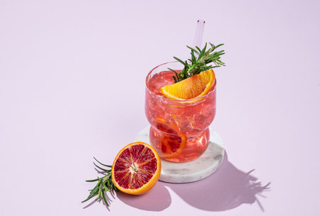 Red orange cocktail with ice and rosemary on a marble stand on a pink background with shadows. Summer citrus drink concept for cafe menu advertising banner. Front view, copy space.の写真素材