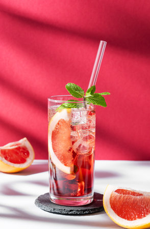 Refreshing cocktail with grapefruit, mint and ice in a glass on a white red background with shadow and sunlight. Summer citrus drink or lemonade concept. Front view and copy space.の写真素材