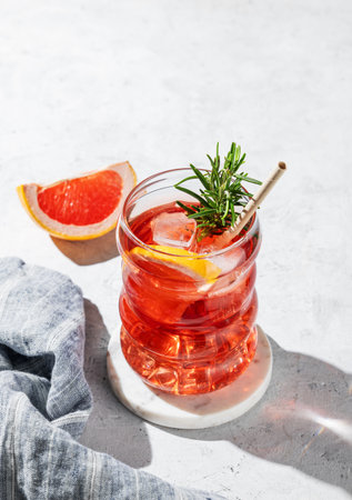 Lemonade with grapefruit, rosemary and ice in a glass on a light background with shadows and napkin. Summer citrus drink or cocktail concept for cafe menu advertising banner. Top view, copy space.の写真素材