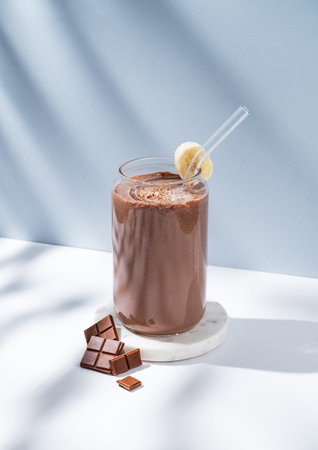 Chocolate smoothie with banana in a glass on a white blue background with morning shadow. Healthy eating and detox diet concept for advertising menu or banner. Front view, copy space.の写真素材