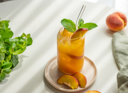 Peach drink or tea with ice, mint in a glass on a white table with fresh fruits, herbs, napkin and sunlight. Refreshing summer cocktail concept for bar or cafe menu. Top view, copy space.の写真素材