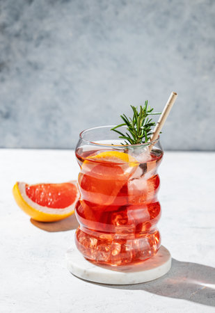 Lemonade with grapefruit, rosemary and ice in a glass on a light background with shadows. Summer citrus drink or cocktail concept for cafe menu advertising banner. Front view, copy space.の写真素材