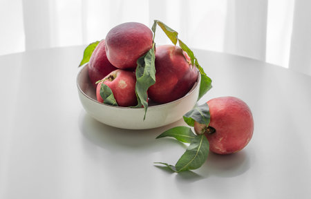 Fresh red peaches or nectarines with green leaves, on bowl on white table and sunlight from window. Healthy and wholesome food concept, organic fruits. Front view, copy space.の写真素材