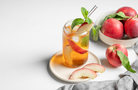 Ice tea with peach, mint in a glass on a white table near window with fresh fruits, napkin and sunlight. Refreshing summer drink concept for bar or cafe menu. Copy space.の写真素材