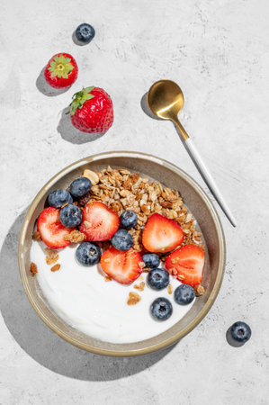 Natural greek yogurt with granola, blueberry and strawberry in a bowl on a light background with fresh berries and shadows. The concept of a healthy, nutritious  breakfast. Top view, copy spaceの写真素材