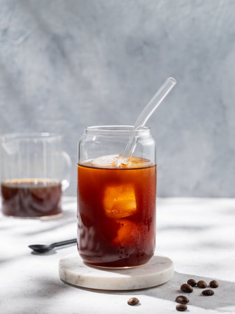Cold brew coffee in a glass with ice on a light background with coffee beans and morning shadows. Trendy summer craft refreshing drink. Front view, copy space.の写真素材