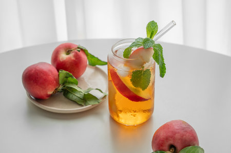 Ice tea with peach, mint in a glass on a white table near window with fresh fruits and sunlight. Refreshing summer drink concept for bar or cafe menu. Front view, copy space.の写真素材