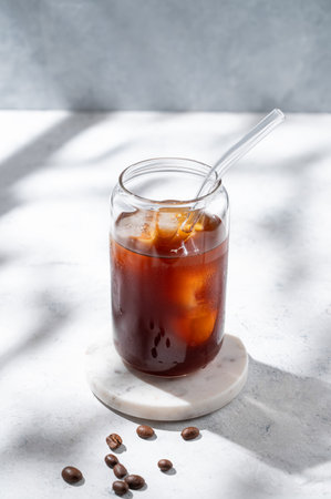 Cold brew coffee in a glass with ice on a light concrete background with coffee beans and morning shadows. Trendy summer craft refreshing drink. Copy space.の写真素材