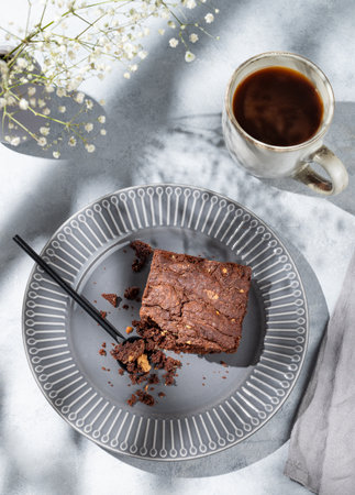 Chocolate brownie with nuts on a dark plate on a light background with cup of coffee, bouquet of flowers and morning shadows. Homemade sweet baking concept. Top view, copy space.の写真素材