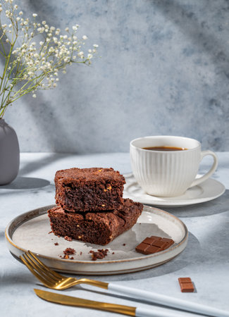 Chocolate brownie with nuts on a plate on a light background with cup of coffee, bouquet of flowers and morning shadows. Homemade sweet baking concept. Front view, copy space.の写真素材