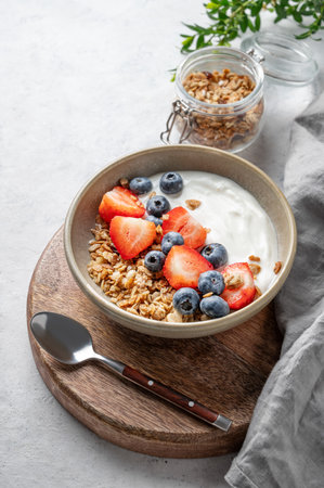 Natural yogurt with granola, blueberry and strawberry in a bowl on a wooden board on a light background with fresh berries. The concept of a healthy, nutritious and dietary breakfast. Copy space.の写真素材